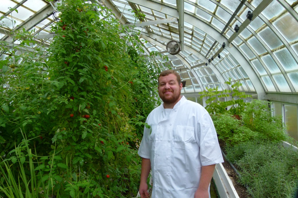 Chef Seth Bailey in The Frick greenhouse next to tomato plants and happy that he has fresh local tomatoes year round.