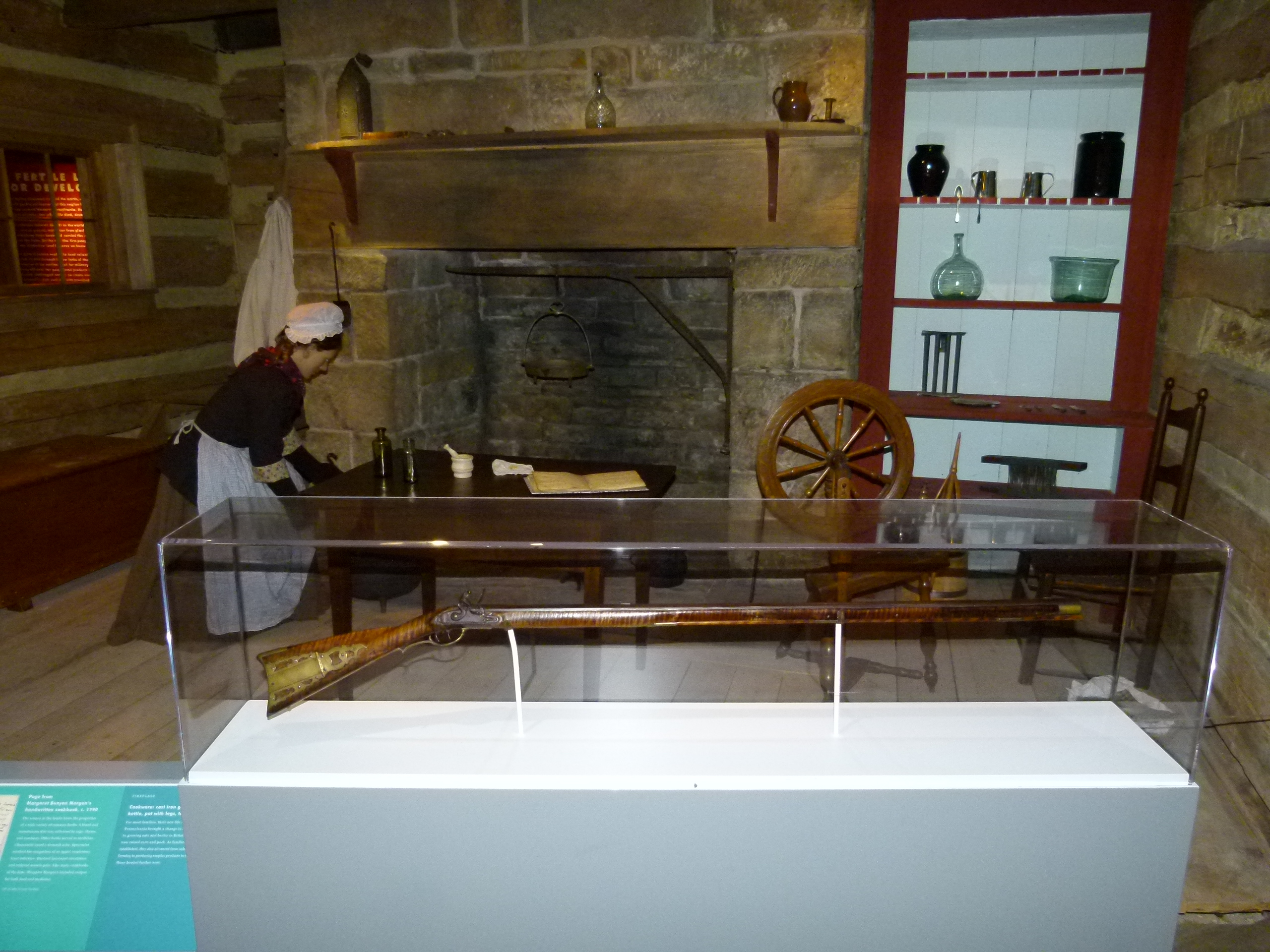 An early American home interior with a hearth, spinning wheel, and long gun.