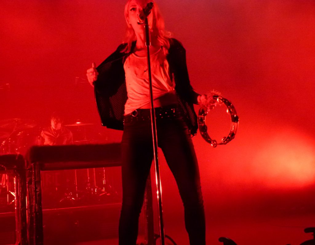 Emily Haines, bathed in red light, does a dance moving while singing and playing the tambourine.