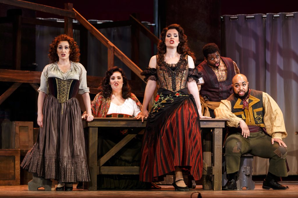 The smugglers, Remendado (Andrew Turner, standing behind the table) and Dancaïre (Yazid Gray), try to enlist the help of the women because of their treachery and thievery. L to R: Frasquita (Véronique Filloux), Mercédès (Leah Heater), and Carmen.