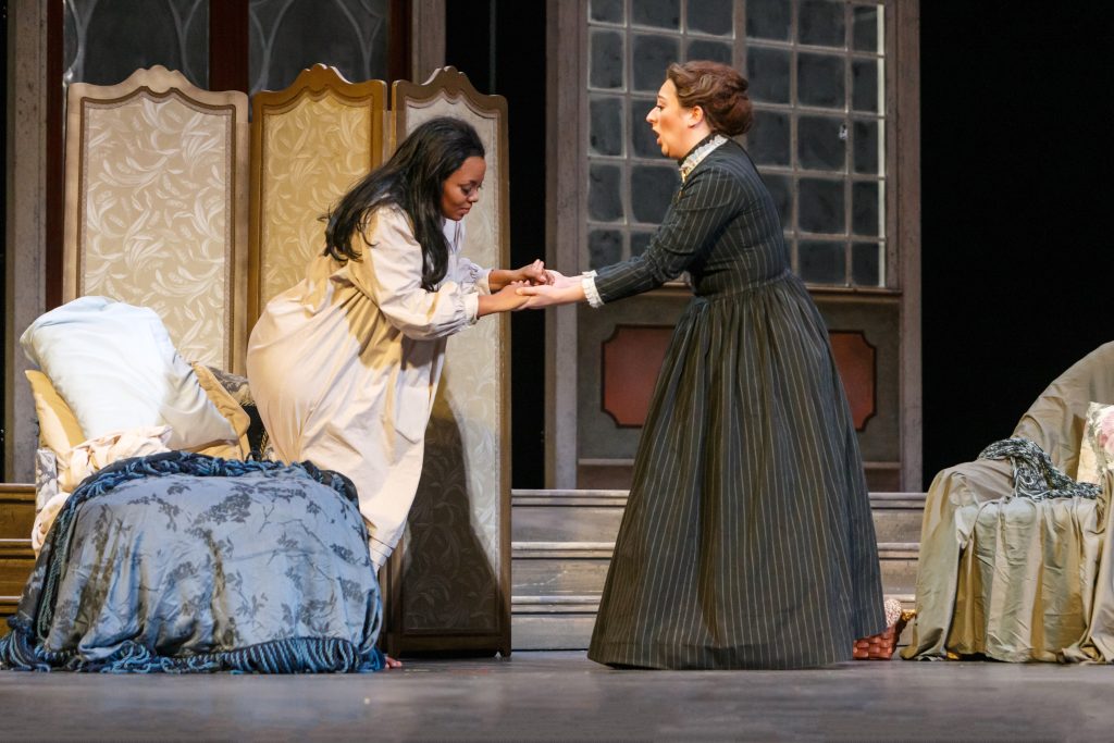 Annina (Julia Swan Laird), ever attendant to her mistress, Violetta Valéry, struggles to get her up from her sick bed.