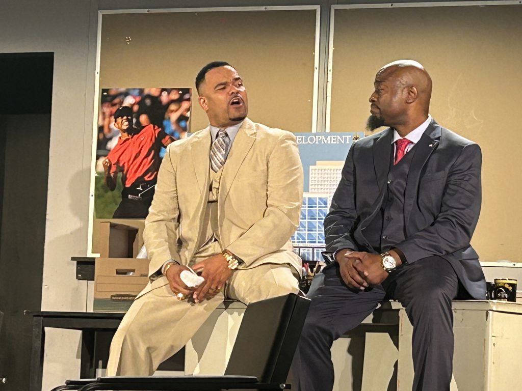 Partner and Tiger Woods fan Hicks (Rich Dickson) informs associate and mayoral candidate Harmond Wilks (Roosevelt Watts, Jr.) how important the game of golf can be in business.