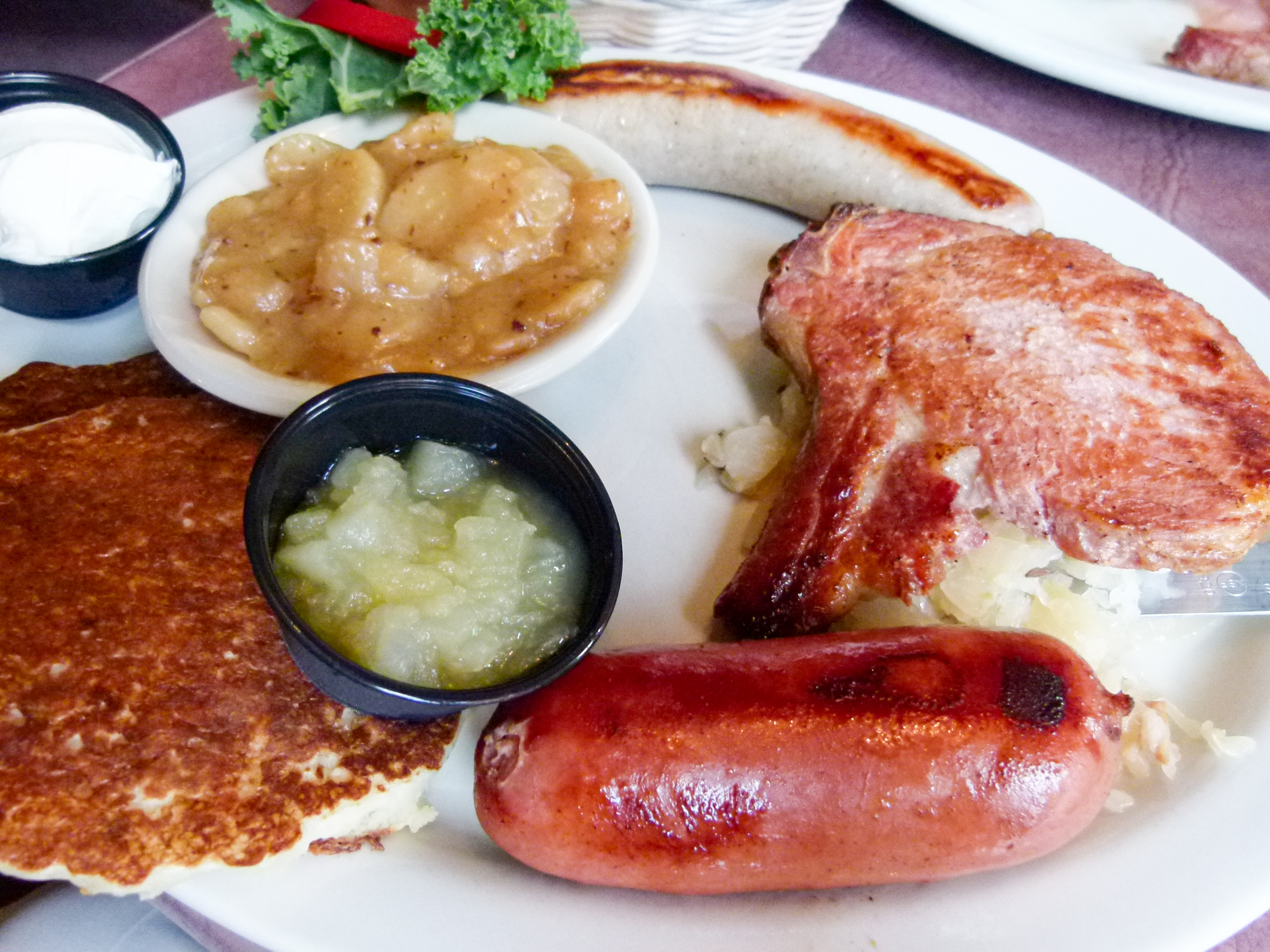 The Bavarian Peasant Platter with (top to bottom) grilled bratwurst, smoked pork loin chop, and knackwurst accompanied by German potato salad, and potato pancakes with apple sauce and sour cream.